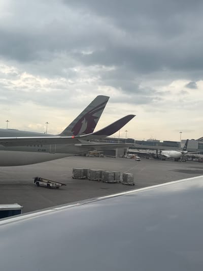 The image depicts an airport tarmac with several airplanes and ground support equipment under a cloudy sky. In the foreground, the wing of an aircraft is visible, with a portion of its fuselage and tail fin extending upwards. The tail fin displays a burgundy and white logo, consistent with Qatar Airways. The registration number "A7-AOB" is visible on the aircraft's fuselage. Further into the scene, another airplane, a white Boeing 777-300ER with the text "ALLIANCE" on its fuselage and tail, is parked at a gate. Ground support equipment, including baggage carts and a tow tractor, are scattered across the tarmac. In the background, airport terminals and structures are visible. The scene is consistent with an airport environment like Toronto Pearson International Airport, located in Mississauga, Ontario, Canada, which serves as a major hub for international flights, including those operated by Qatar Airways. There are no people visible in the image.