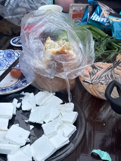 The image depicts a table in what appears to be an outdoor setting, possibly in Sukhobuzimskoye, Russia. A black plate is prominently displayed, filled with cubed white cheese. A plastic bag containing bread sits near the plate, alongside a bowl with a swirled tan and white mixture. Fresh green herbs or vegetables are present. A patterned plate with remnants of food is visible. Other items, including packaging, are also present on the table. The scene appears to be a casual meal setting.