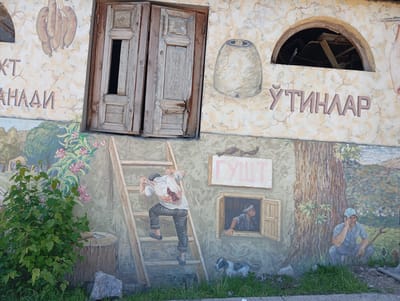 A mural adorns a building in Chinarkent, Uzbekistan. The mural is situated on a wall, above the ground level, featuring both painted and physically incorporated elements.
On the upper portion of the mural, a pair of old wooden doors, seemingly mounted on the wall, appears slightly askew, with one side open. Above the doors, images of sausages and a rounded oven are painted. An open arch or hole is present on the upper right corner of the wall. In this section of the mural, lettering in a different language is visible.
The lower portion of the mural depicts a scene with painted figures and objects. A man is shown climbing a ladder, carrying a butchered animal over his shoulder. Another man looks out from a window located on the lower right. A dog sits at the base of the wall. A man with a cap is squatting with a shovel and on a cellphone in front of a large tree. In the lower left, there is a depiction of greenery and a tree stump.
The overall scene is a combination of painted and three-dimensional elements, giving the mural a unique and somewhat surreal quality. The presence of text in a different language, the depiction of rural life, and the location in Chinarkent, Uzbekistan, provide cultural context to the image.