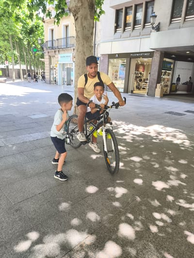 In Vitoria-Gasteiz, Spain, a man wearing a yellow t-shirt and a black baseball cap is riding a bicycle with two young boys. The man is seated on the bicycle, with one boy sitting in front of him and another boy standing on the ground beside the bicycle, looking up at the man and the boy on the bike. The boy in front is smiling and making a peace sign with his fingers. The scene takes place on a paved sidewalk with dappled sunlight filtering through the trees overhead. In the background, there are buildings with shopfronts, including one with the name "PACOMARTINEZ" visible. The overall impression is a family outing on a sunny day in a city environment.