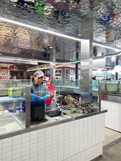 A restaurant worker in blue gloves stands behind a glass-enclosed oyster display, washing oysters under a running faucet. The display is stocked with numerous oysters. Behind him, another person in a red shirt is visible. The interior features a reflective, wavy ceiling and tiled walls. "It's HOTBOYS" signage hangs in the background, indicating the restaurant's name. Boxes and supplies are seen on the oyster preparation area.