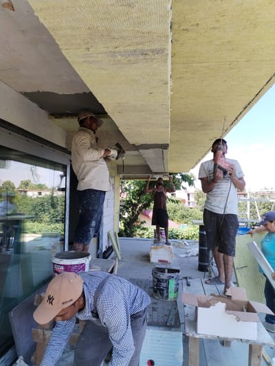 The image shows a group of construction workers on a balcony, likely in Dumbrăveni, Romania. The scene is outdoors, with a clear blue sky and lush greenery visible in the background, suggesting a sunny day. The workers are engaged in construction or renovation work, focusing on insulating the exterior of the building.

Several individuals are actively participating in the task. One man, wearing a light-colored shirt and a cap, is standing near a large window and appears to be applying something to the ceiling with a trowel. Another man, in a brown tank top and dark shorts, is standing on a makeshift platform and holding his arms up, possibly in a supportive role or signaling. A third worker, wearing a light blue t-shirt and dark shorts, is holding a red power drill up to the ceiling, suggesting he is drilling holes for installation. A fourth person, wearing a blue t-shirt and a cap, is visible on the right, holding a large insulation panel. In the foreground, a person wearing a checkered shirt and a New York Yankees baseball cap is bent over, seemingly working on something on the floor.

Various construction materials and tools are scattered around the area, including buckets, cardboard boxes, insulation panels, and a hammer. One bucket has the text "KRAFT PAINTS" visible on it. The overall impression is one of active construction work on a bright day, with the workers focused on their tasks.