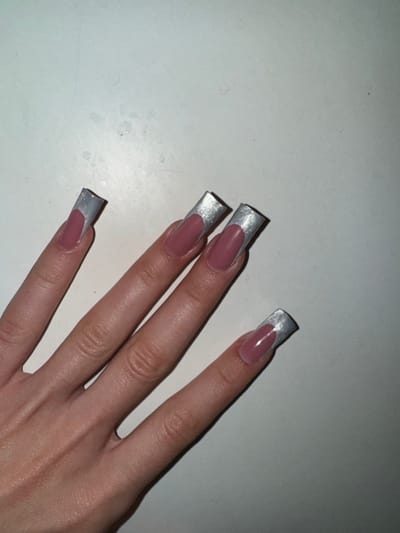 A person's hand with four long, square-shaped artificial nails is visible against a light-colored, textured surface. The nails are decorated with a pink base color and a metallic silver French tip. The hand is positioned diagonally from the bottom left to the top right of the frame. The environment appears to be indoors, with the surface possibly being a desk or tabletop. No specific details about the location in Сароцкі сельскі Савет, Belarus are discernible from the image.