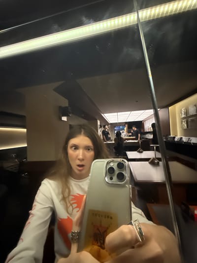 A young woman with wide eyes and an open mouth takes a selfie in a mirror. She is wearing a white long-sleeved shirt with an orange graphic design. She has two silver rings on her fingers. In the background, reflected in the mirror, is a bar or restaurant with several people working behind the counter and patrons seated at tables. The scene appears to be indoors, possibly at night, with artificial lighting. The reflection also shows a long fluorescent light fixture above the main dining area. The overall impression is that of a casual, possibly dimly lit, establishment. The location is not specified as being in Jing'an District, China.