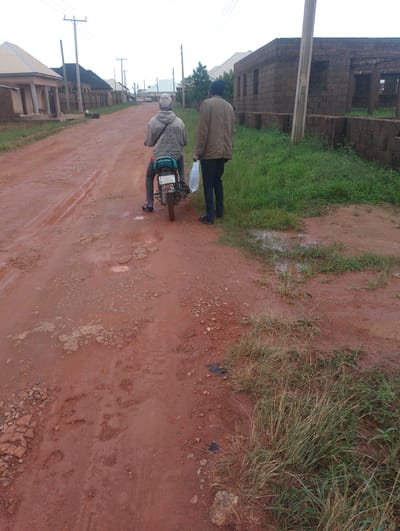 This is a medium shot, eye-level, outdoor photograph taken on a reddish-brown dirt road in Russo, Nigeria. The road is uneven with several visible potholes and puddles, suggesting recent rainfall. On the left side of the road, there are several residential buildings with light-colored walls and dark roofs. Beyond these, a row of fence posts is visible. Further down the road, the outlines of more buildings and utility poles can be seen against an overcast sky.

In the foreground, two men are present. The man on the left is seated on a motorcycle, facing away from the camera. He is wearing a grey hooded sweatshirt with text on the back, dark pants, and black sandals. The motorcycle appears to be a light-colored model with a blue bag strapped to the back. The man on the right is walking beside the motorcycle, also facing away from the camera. He is wearing a brown jacket, dark pants, and black shoes. He is carrying a clear plastic bag in his left hand.

To the right of the road, there is tall, green grass and some patches of bare, reddish-brown earth. In the mid-ground, a partially constructed building made of dark brick is visible. A concrete utility pole stands in front of this building. The overall scene depicts a typical streetscape in a developing urban or semi-urban area, characterized by unpaved roads, modest housing, and ongoing construction.