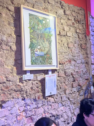 The image depicts an interior scene within an establishment in Bogota, Capital District, Colombia, characterized by a prominently featured, rustic, exposed brick wall. A large, vertically oriented artwork is mounted on the wall, enclosed in a light-colored wooden frame. The artwork illustrates a detailed jungle environment with a brown adult sloth hanging from a branch, cradling a smaller, lighter-colored baby sloth. Below the framed art, a small white label provides item details including a price in COP (Colombian Pesos), and to its right, a larger white poster is affixed, bearing the red title "FUERZA DE DOS SERES: EL TIEMPO DEL BOSQUE" (Strength of Two Beings: The Time of the Forest), accompanied by a block of Spanish text and a black and white symbol. The brick wall features visible mortar and varies in color from reddish-brown to grey, with some areas showing signs of past plaster. A strip of red paint is visible along the top edge of the wall, and a red electrical conduit runs vertically on the right side. The lower portion of the frame shows the partial heads and shoulders of at least two individuals, one wearing glasses, indicating seated patrons or visitors in the foreground. The scene is illuminated by artificial light, casting subtle shadows and creating a slight purple hue on the lower brickwork.