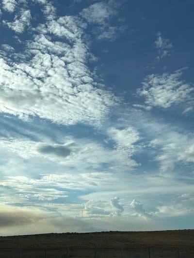 A wide shot captures a vast expanse of sky above a gently rolling, dry, grassy landscape in the city of Төртқұдық, Kazakhstan. The sky is filled with a variety of clouds. In the upper portion of the frame, altocumulus clouds create a textured, rippled pattern against a deep blue sky. Lower down, cirrus and stratus clouds stretch across the horizon, interspersed with patches of lighter blue sky. Towards the left of the frame, a plume of smoke or dust rises from the horizon, tinged with orange and brown hues, suggesting potential smoke from a fire or a dust storm. The foreground consists of a flat, open field of dry, light brown grass, with a few horizontal wooden fence posts visible. The lighting suggests it is daytime, with the sun likely positioned off-frame, casting a soft, diffused light across the scene. There are no people or animals visible in the image.