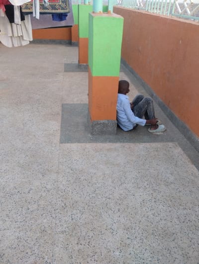 A person sits against a pillar on a rooftop. The pillar is painted green on the top half and orange on the bottom. The person wears a light blue shirt and grey patterned pants. Their legs are bent. They are wearing light-colored shoes. An orange wall extends along the right side of the pillar. The rooftop is a speckled, light-colored surface. There are more pillars in the background with clothes hanging on a clothesline to the left. A white metal fence is visible in the far background.