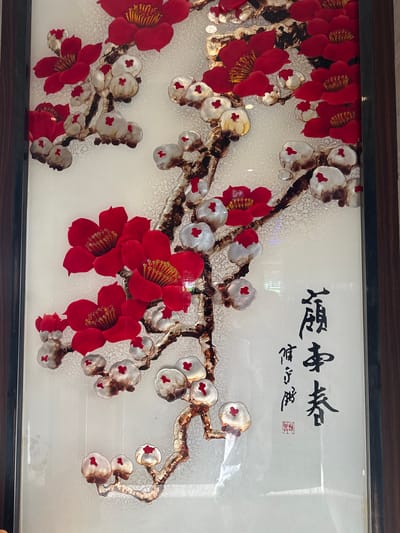 This is a close-up image of a framed piece of art, likely a decorative wall hanging. The artwork depicts a branch with blooming flowers, primarily in vibrant red and white with gold accents. The flowers appear to be made of a textured material, possibly fabric or paper, giving them a three-dimensional effect. The branch itself is rendered in dark brown and gold, suggesting a naturalistic yet stylized representation. The background is a light, textured white, which makes the floral elements stand out.
In the upper right portion of the artwork, there is visible East Asian calligraphy in black ink. The characters are flowing and artistic. Below the calligraphy, a small red seal with intricate markings is present, common in East Asian art to denote ownership or authenticity.
The framing is dark wood, giving the piece a finished and elegant look. The image is taken indoors, as suggested by the lighting and reflections on the glass cover of the artwork. There are no people or specific environmental details like weather or time of day. The overall impression is one of artistic beauty and cultural significance, likely intended for interior decoration.