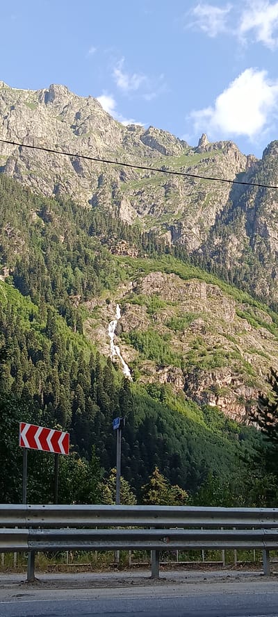 The image shows a mountainous landscape with a waterfall cascading down a rocky slope. Lush green trees cover the lower part of the mountain, transitioning to rocky, sparse vegetation higher up. In the foreground, a road barrier is visible, along with a chevron sign indicating a sharp curve to the left. A tall pole with a blue sign is also present. Power lines stretch across the upper portion of the image against a bright blue sky with scattered white clouds. This scene is consistent with the natural scenery found in Dombay, Russia, known for its Caucasian mountain range and natural attractions like waterfalls.