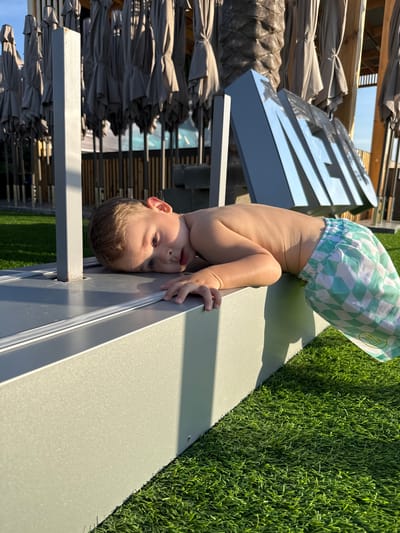 A young boy with light brown hair is lying on his stomach, resting his head on a metallic surface. He is wearing checkered shorts. The boy's left hand is visible, resting on the metallic surface. In the background, there are many closed umbrellas, a palm tree trunk, and a large metallic sign with letters. The scene is outdoors, with artificial grass on the ground. The image appears to be taken in a recreational area, possibly a beach or resort, located in the city of Domino, Russia.