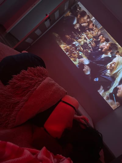 A person is lying on a bed in a dimly lit room, covered by a fuzzy blanket and a red blanket. A projector is casting an image onto a wall, showing a scene of people gathered around a long table. The person on the bed appears to be watching the projected image. The room is decorated with red lighting. There is no discernible connection to Gliwice, Poland in the image.