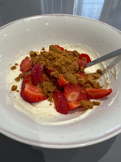 A close-up, eye-level shot shows a white bowl filled with a dessert of yogurt, sliced strawberries, and crumbled biscuits. The bowl has a light pink rim and a speckled texture. A silver spoon is partially visible on the right side of the bowl, resting in the dessert. The background is a dark grey surface. The image is well-lit, with a slight glare on the yogurt. There is no information in the image to indicate the location in Craigavon, United Kingdom.