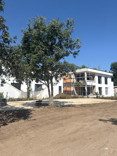 A contemporary, unfinished white house with a flat roof is visible under a clear blue sky. Scaffolding surrounds a section of the building where exposed brickwork is present, suggesting ongoing construction. Several windows, some covered and some clear, are distributed across the structure. In the foreground, a large, leafy tree with green fruits stands prominently. The ground in front of the house is largely unlandscaped, consisting of dirt and some scattered construction materials, including wooden pallets and red and white caution tape. Shadows from the tree and other unseen objects are cast across the scene. To the left, a large dark pot with some greenery is visible, and to the right, a palm tree is planted near the house. The overall impression is of a construction site in a rural or suburban setting, possibly in the city of Shabla, Bulgaria, given the context.