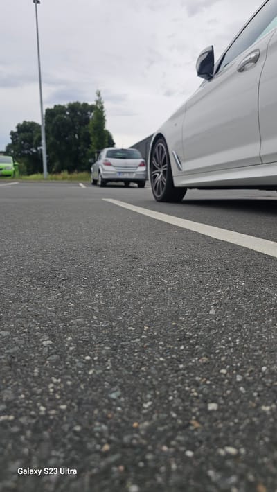 The image depicts an outdoor scene, likely a parking lot, with a low-angle perspective. The foreground is dominated by asphalt, showing a textured surface with visible gravel. A white line is painted on the asphalt, running diagonally across the image from bottom right to top left.
In the middle ground, a silver car is parked. Its rear is visible, and it appears to be an older model hatchback. Further behind the silver car, a tall, thin pole, possibly a street light or utility pole, stands vertically. Trees with dense foliage are visible in the background, suggesting a natural environment adjacent to the parking area.
The right side of the image features the rear portion of a white modern car, including its wheel and part of the door. The car is parked on the asphalt, and its side profile is partially visible.
The sky is overcast, with diffused light indicating a cloudy day.
There are no people or animals visible in the frame. The scene appears to be static, with no discernible action or interaction taking place. The overall impression is that of a quiet parking area under a cloudy sky.
The watermark "Galaxy S23 Ultra" is present in the bottom left corner of the image.
While the image itself does not provide explicit markers of location, the provided context states the scene is in the city of Schleiz, Germany. Given the appearance of the cars and the general environment, this is a plausible setting.