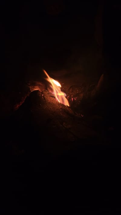 The media file presents a close-up, low-light photograph of a fire burning in a dark environment. The central subject is a vivid flame, glowing brightly with intense orange, yellow, and red colors. The flames appear somewhat blurred, suggesting either natural flickering, slight motion during capture, or a longer exposure time, creating streaks of light.

The fire is situated within or next to a dark, textured surface, most likely a log or piece of wood, which is partially illuminated by the firelight. This surface shows details of its rough texture, possibly bark or charred material, rendered in reddish-brown and dark tones. Scattered around the main flame are smaller, glowing red spots, indicative of embers burning intensely.

The background is predominantly dark black, with very little discernible detail, emphasizing the isolation and brightness of the fire. This suggests the photograph was taken at night or in a very dark setting, where the fire is the primary light source. The overall impression is one of warmth and elemental beauty against the surrounding darkness. Given the location context of Khir Ganga, India, this could be a campfire or a fire for warmth in a natural, possibly mountainous or trekking environment. There are no people, other distinct objects, or visible text present in the image.