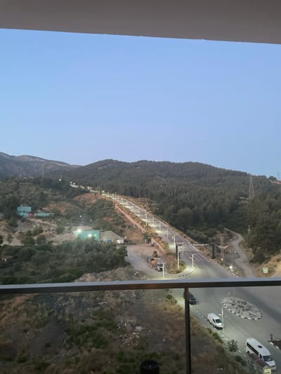 The image captures a view of Yunus Emre Culture Park in Yunus Emre, Turkey, as dusk settles. A road, illuminated by streetlights, winds its way up a hillside covered in trees. The park entrance, marked by an archway with Turkish writing, is visible along the road. On the left side of the road, there are several green buildings, one of which has a Turkish flag. Construction equipment, including a bulldozer, is parked near the buildings. Further down the road, vehicles, including vans and cars, are present. The landscape is characterized by rolling hills and dense forestation. The sky is a clear, pale blue, with a faint hint of the moon. In the foreground, a railing suggests the vantage point is from a balcony or elevated structure.