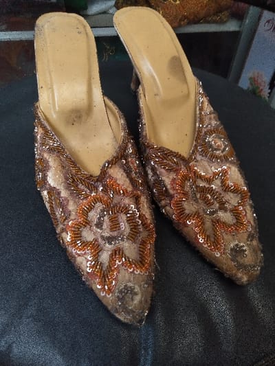 A pair of worn, embellished women's slip-on high-heeled shoes, commonly known as mules, rests on a dark, textured surface. The shoes feature a light tan or beige fabric base heavily adorned with intricate floral patterns composed of small, reddish-orange and bronze-toned beads and sequins. The pointed toes show signs of scuffing and discoloration. The interior soles are light tan, exhibiting visible dark smudges and dirt accumulation in the heel and arch areas, indicative of prior use. The heels, partially obscured, appear to be of moderate height. In the blurred background, hints of other decorative items, possibly a similar embellished object and a partially visible book or frame with a floral motif, suggest an indoor setting. There are no individuals present. No city or country information is discernible from the image content.