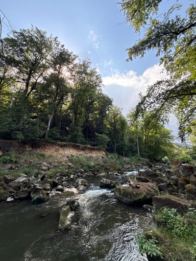 A person is sitting on a large rock next to a flowing river. The person is wearing dark shorts, a light-colored t-shirt, and has a red backpack next to them. The river is surrounded by mossy rocks and lush green trees. The scene is shot from a high angle, looking down on the river and the person.

The sun is shining through the trees, creating dappled light and casting shadows on the ground. The sky is blue with a few wispy clouds. The water is dark and appears to be moving quickly, with some white foam visible. The rocks are uneven and appear to be part of a natural landscape, possibly a gorge or a waterfall.

The overall impression is a peaceful and serene scene, with the person enjoying the natural beauty of the surroundings. The time of day appears to be late afternoon, given the angle of the sun. The location cues, such as the rocks, trees, and river, suggest a natural, outdoor setting. There is no visible text in the image.