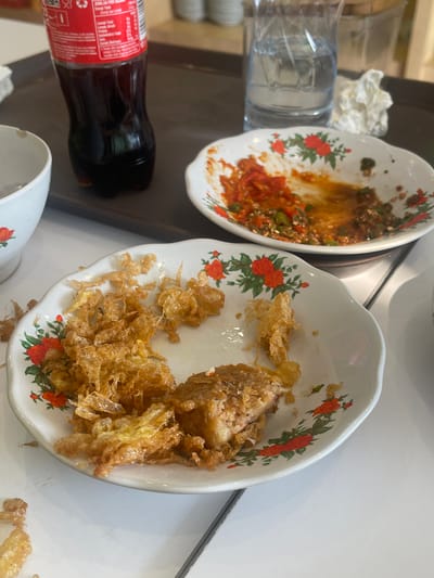 The image shows a close-up view of remnants of a meal on a white table. In the foreground, a white, floral-patterned plate contains a portion of fried food, possibly fish or meat, along with crispy fried batter. A smaller, similar plate in the background holds leftover chili sauce and chopped green chilies. A partially full glass of water sits behind the plates, next to a crumpled white napkin. A dark bottle of Coca-Cola is visible to the left. The overall impression is that of a meal that has recently been consumed. The image does not provide any specific information about the location being Medang, Indonesia.
