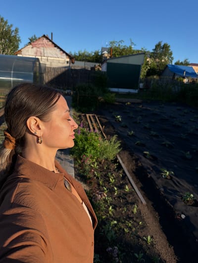 The image depicts a profile view of a woman in the foreground, facing right with her eyes closed. She has brown hair pulled back into a ponytail, wears a brown collared shirt, and an earring in her left ear. Behind her, a garden plot is visible with rows of plants covered by black fabric. Several structures, possibly houses or sheds, are present in the background, along with trees and a bright blue sky. The scene appears to be a residential area with a garden. The overall lighting suggests it is either early morning or late afternoon. The location is Kansk, Russia.