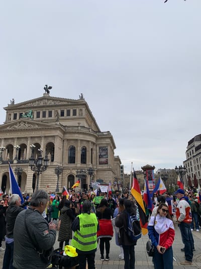The image shows a group of people gathered in front of the Alte Oper, an opera house located in Frankfurt am Main, Germany, at the coordinates 50.115 latitude and 8.672 longitude. The people are holding signs and flags, and some are wearing red and yellow vests. There are multiple flags, including the Philippine flag, being displayed. The flags are held in the air by the protesters. The opera house is a large, ornate building with a statue of a winged horse on its roof. It is a beautiful and historical building. The sky is cloudy, but the image is still bright. The atmosphere is upbeat and cheerful, despite the protest. The protesters appear to be celebrating the Philippine flag and enjoying themselves. The focus of the photo is on the people and the flags. The opera house is a backdrop.