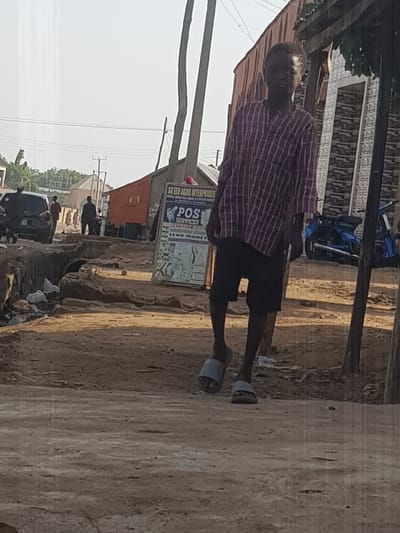 A full-body shot of a young Black male is captured in an outdoor setting, likely a street in Kufena, Nigeria. The individual is wearing a red and white plaid long-sleeved shirt and black shorts, with gray slide sandals on his feet. He is walking towards the viewer, with his head slightly bowed and his gaze directed downwards. His posture suggests a casual, perhaps pensive, demeanor.
The environment appears to be a dirt road with uneven terrain, characterized by loose soil, rocks, and some scattered debris, including plastic waste on the left side of the frame. In the background, several structures are visible. To the left, a dark-colored car is parked, and a few other individuals are present, though less distinct. A red cargo truck is also visible, with a sign on its side displaying "POS SERVICES," "DEPOSIT MONEY," and "SEND MONEY." Behind these elements, a building with a textured facade and what looks like a metallic roof is present. The sky is bright, suggesting daytime, with a slightly hazy or overcast condition. Utility poles and overhead wires are also part of the background landscape, indicating an inhabited area. The overall impression is that of a typical street scene in a town or city.