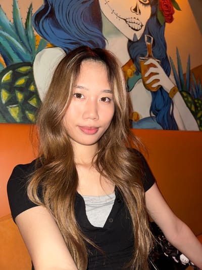 A medium shot captures a young East Asian woman posing indoors, her body angled and head turned towards the camera. She has long, wavy, light brown hair with prominent blonde highlights, cascading over her shoulders and chest. Her eyes are open, and she gazes directly at the viewer with a calm, slightly smiling expression, her lips naturally colored and slightly parted. She is wearing a black cardigan or button-down top over a grey inner shirt. Her right arm is visible, extended towards the top right of the frame.
The background features a vibrant mural on a light-colored wall, depicting a stylized female figure. The figure has dark blue hair adorned with a red rose and a face styled like a calavera or sugar skull, with prominent cheekbones and a black, leaf-like motif on her cheek. One hand of the painted figure is holding a small, amber-colored bottle. Below the figure, there are elements resembling green pineapple or agave leaves. To the right of the mural, a plain orange wall or surface is visible, providing a contrasting warm tone. The lighting suggests an indoor setting, possibly an evening or night, with a warm ambient glow. No text is visible in the image.