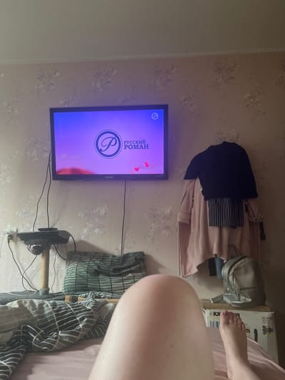 The image shows a bedroom scene, likely in Tula, Russia, with a television displaying the title "РУССКИЙ РОМАН" (Russian Romance) and a prominent stylized letter "P" within a circle. The television is mounted on the wall, and beneath it, a black object with fuzzy texture and a scratching post is visible, connected to electrical outlets. Various clothing items, including a dark blue shirt and a pink robe, are draped over furniture or furniture-like surfaces, suggesting a lived-in space. A silver backpack sits on top of a cardboard box. In the foreground, a person's foot with purple nail polish is visible, resting on a pink surface, possibly a bed or blanket. The room appears to be dimly lit, suggesting it could be evening or nighttime. The wallpaper has a floral pattern. The overall atmosphere is casual and domestic.
