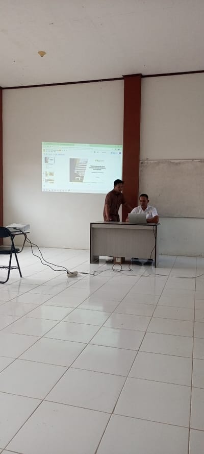 Two men stand in a tiled room. One, in a brown shirt, leans toward the other, who is seated at a desk with a laptop. A presentation is projected on the wall behind them. A projector sits on a chair to the left, connected to a power strip on the floor with visible wires. A brown pillar and a whiteboard are visible against the white walls.