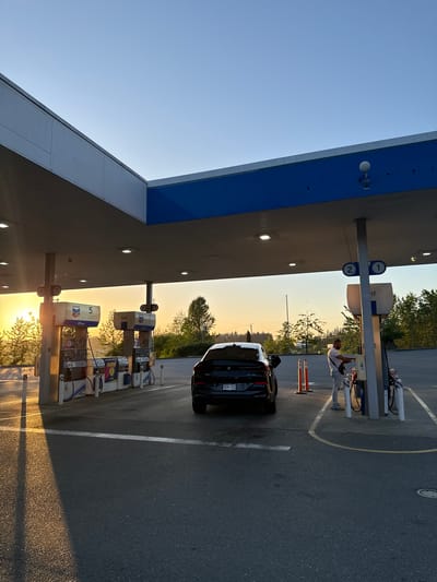 The photograph depicts a gas station in Parksville, Canada. A black car is parked at pump number 2. A person is standing near the pump on the driver's side of the car. The sun is low on the horizon, creating long shadows and a warm light. Several other gas pumps are visible in a row. The sky is clear and blue. The gas station canopy has blue accents.
