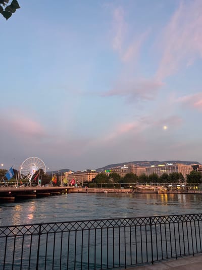 This image captures a waterfront scene in Geneva, Switzerland, at dusk. The sky is a soft gradient of pale blue and pink, with a faint moon visible. A calm body of water, likely a river or lake, reflects the lights from the buildings lining the shore.

Along the waterfront, there's a promenade with trees and buildings. Some of the buildings display illuminated signage, including "Cartier," "J.P. Morgan," "Patek Philippe," and "Rolex." This suggests a high-end commercial area. People can be seen walking along the promenade and seated at outdoor cafes or restaurants.

A Ferris wheel, illuminated with white lights, is a prominent object in the scene, suggesting a possible fair or festival is taking place. Several flags are also visible, including Swiss flags. A black metal railing runs along the right side of the image, separating the viewer from the water. The overall atmosphere appears peaceful and lively, with the warm glow of lights contrasting with the twilight sky. The presence of the Ferris wheel and flags suggests a festive occasion, while the brand names on the buildings hint at a sophisticated urban environment.