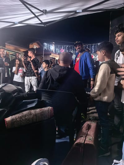 In the city of Rozino, Bulgaria, a gathering of people is observed outdoors at night, under a white canopy supported by metal poles. A group of individuals are visible, some interacting with each other. In the foreground, a dark suitcase with metal latches and handles is positioned horizontally, with a patterned textile object placed on top of it. Behind the suitcase, a dark chair is occupied by a person seen from the back, facing away from the camera. Further back, a man in a blue jacket, a red shirt, and a gold chain is interacting with others, and appears to be wearing a cap and sunglasses. To his right, a young person in a cream-colored fleece jacket and jeans is looking towards the man. Several other people are visible in the background, including a person holding a smartphone, and others standing and looking towards the main group. Decorative string lights are illuminated on buildings in the background, suggesting a festive or celebratory occasion. The overall scene depicts a social event taking place in an outdoor urban or suburban setting at night.