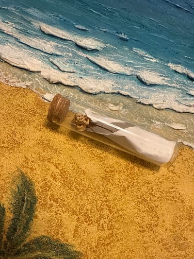 A glass vial with a cork stopper lies on a textured sandy surface. Inside the vial, a small, dried flower and a rolled piece of paper are visible. The background depicts a textured representation of ocean waves crashing onto a beach. The image is taken from a high-angle perspective. There is no indication of the city of Drozhzhino, Russia, in this image.