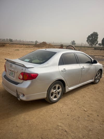 A silver Toyota Corolla sedan, bearing Nigerian license plate "ABJ 355C," is stationary on an unpaved, reddish-brown dirt surface in Gyellesu, Nigeria. The vehicle is angled with its rear and right side visible. It features a rear spoiler, tinted windows, and alloy wheels. The lower left section of the rear bumper exhibits minor damage, including a scuff and deformation. The rear left quarter panel shows scuff marks on the paint. The ground is dry and dusty. In the background, multiple mounds of earth are present, suggesting an excavation or construction site. Partially visible construction equipment, possibly a concrete mixer, is located behind the vehicle's midsection. Sparse trees with green foliage are scattered across the distant, hazy landscape under a pale, overcast sky. No individuals or active interactions are visible.