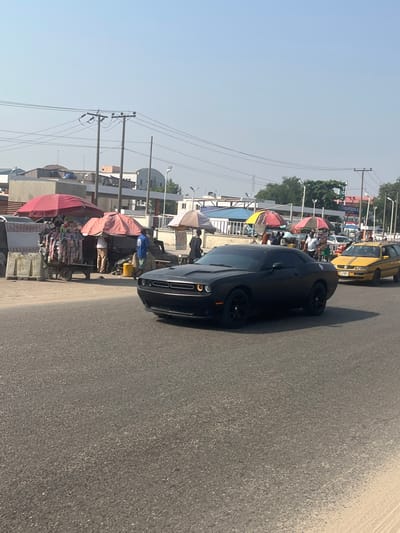 The image captures a street scene in Ikota, Nigeria, during daylight hours under a clear, light blue sky. A matte black Dodge Challenger is positioned prominently in the central foreground, moving from right to left across an asphalt road, with its headlights illuminated.
To the left of the road, a series of street vendors operate under various colored umbrellas, including red, pink, orange, and beige. One vendor's cart on the far left displays clothing items. Several individuals are present near these stalls; one person is seen wearing a light blue shirt, another a dark shirt. Behind the initial vendor setups, large concrete barriers or blocks are visible. Utility poles with multiple overhead power lines crisscross the upper-left portion of the frame, with distant buildings, including a multi-story structure, discernible in the background.
To the right and slightly behind the Challenger, a yellow vehicle, possibly a station wagon or sedan with dark side stripes, is visible on the road. Further down the street on the right, additional vendor stalls with umbrellas, pedestrians, streetlights, and a sign partially legible as "GAPDEN C" are present. The overall environment depicts an active urban or semi-urban thoroughfare characterized by vehicular traffic and roadside commercial activity.