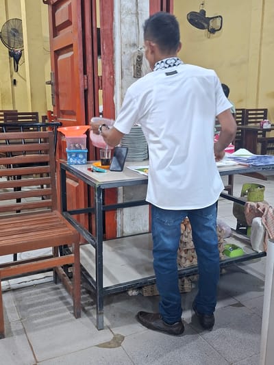 The image shows a person in a white shirt and blue jeans standing at a table in what appears to be a food establishment in Keudegeudong, Indonesia. The person is holding a plastic container and pouring something into a glass. On the table, there is a smartphone, a lighter, a stack of plates, and some papers. In the background, there are wooden chairs and tables, a red door, a wall-mounted fan, and some stacked items including egg cartons. The lighting suggests an indoor setting, possibly a warung or small restaurant.