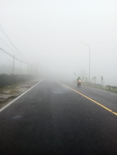 The image portrays a fog-obscured asphalt road in Dawathang_Dorjibi_ Kashingtsawa, Bhutan. Dense white fog restricts visibility significantly, creating a uniform light grey background. The road surface is dark and wet, indicating moisture. A continuous white line marks the left edge of the road, while a continuous yellow line defines the center on the right side. On the left, a dark, wet dirt shoulder runs parallel to a metal fence, behind which several utility poles with multiple power lines extend into the fog. On the right, a metal guardrail is visible, with sparse, low-lying vegetation beyond it. Multiple streetlights stand along this side, their upper portions obscured by the fog. A single individual, dressed in a light green top, is operating a dark-colored motorcycle. The motorcycle's headlight is illuminated, and the vehicle is positioned on the right side of the road, moving away from the viewer.