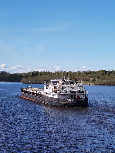 The image depicts a black and white barge labeled "КУНЦЕВО" (KUNTSEVO) on a wide body of water, likely a river. The barge is oriented away from the viewer, moving through the water and creating a small wake. The river is flanked by a tree-lined bank and a clear blue sky with scattered clouds.
The barge is in Pruzhinino, Russia.