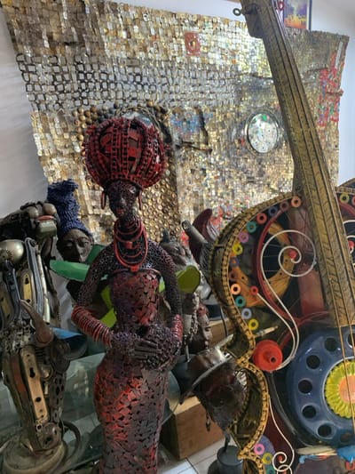 The scene, an interior art display located in Ikate, Nigeria, features an array of sculptures crafted from repurposed materials. A prominent, tall female sculpture, constructed from interconnected dark red metal components, stands centrally. This figure displays an elaborate crown, multi-strand necklaces, and bangles, all consistent in material and dark red hue. To her immediate left, a partially visible, abstract humanoid sculpture is present, fashioned from various metallic parts and topped with a blue knitted headpiece. The background is dominated by a large wall mosaic composed of numerous flattened metallic rectangles, likely recycled can materials, creating a textured, shimmering surface. This wall art includes irregular dark patches and a circular element containing embedded compact discs. To the right, a large, stylized string instrument, resembling a cello or double bass, is visible. This instrument is assembled from metal, bicycle chains, gears, and colorful rings, with a blue telephone dial integrated into its body. A smaller, dark-toned head sculpture wearing a fedora-style hat is partially visible at the base of the string instrument. Sections of white wall are visible on the left and in the upper background.