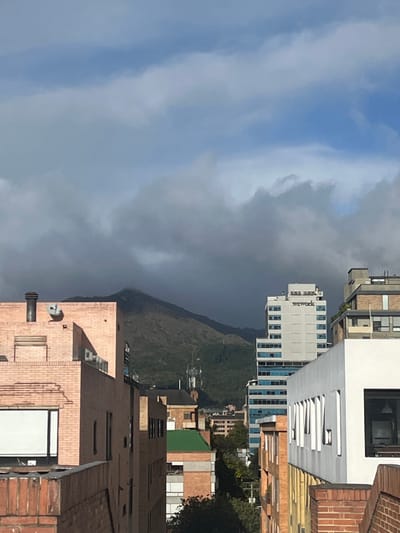 The image depicts an urban cityscape in Bogota, Capital District, Colombia, with a prominent mountainous backdrop under a partially clouded sky.

The scene is dominated by a large, vegetated mountain in the central background, primarily covered by dense, dark gray clouds that obscure its peak. Portions of the mountain's lower and mid-slopes are visible, showing green foliage and several communication towers or antennas. Above the dark clouds, patches of lighter blue sky are visible, mixed with white and light gray stratiform clouds.

In the midground, several buildings are visible. On the right, a tall, multi-story, light gray or white skyscraper features the "wework" logo prominently near its top. This building has numerous horizontal bands of blue-tinted windows. Adjacent to its right is a darker, flat-roofed building. In the immediate right foreground, a section of a white, modern building with distinct recessed rectangular window openings is present, below which a reddish-brown brick wall or structure is visible.

On the left midground, a light reddish-brown brick building with a dark, cylindrical chimney-like structure and a small, light-colored satellite dish on its roof is visible. Below its roofline, a white-framed window is partially seen. Further back and to the right of this brick building are other structures, including one with a green-sloped roof and others of various heights and materials, indicating a dense urban environment.

No individuals, vehicles, or specific actions are discernible in the frame. The lighting suggests daylight, possibly with the sun casting shadows from the right.

The visible "wework" building at Carrera 13 #93A-67, Parque 93, Bogota, identifies the specific location within the city, with the mountain in the background being part of the Cerros Orientales.