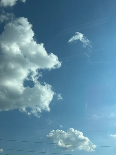 This media file captures a clear blue sky with scattered white, puffy cumulus clouds. The perspective is upward-looking, suggesting the photo was taken from the ground or a low vantage point. Two thin, dark, vertical lines are visible on the right side of the image, likely power lines or wires, indicating an outdoor, potentially semi-urban or rural setting. The sunlight suggests it is daytime, likely a bright, clear day. There are some faint reflections or lens flares visible across the sky, particularly a diagonal streak on the left side and a more subtle one on the right. The clouds are dynamic in shape and texture, with some appearing wispy and others more dense and voluminous. The overall impression is one of a serene and bright day. There is no visible text in the image.