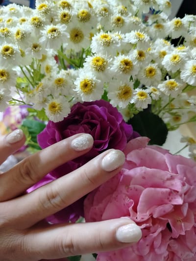 The image shows a close-up view of a person's hand with manicured nails in front of a bouquet of flowers. The bouquet consists of white flowers with yellow centers, possibly chamomile or a similar daisy-like flower, and two large roses, one deep purple and one pink. The hand is positioned to the left of the frame, with fingers gently touching one of the flowers. The fingernails are painted a light, pearlescent white with subtle floral designs on some. The background is filled with the dense arrangement of flowers, creating a soft and colorful backdrop. There is no indication of the location being in Vazerki, Russia, or any specific city.