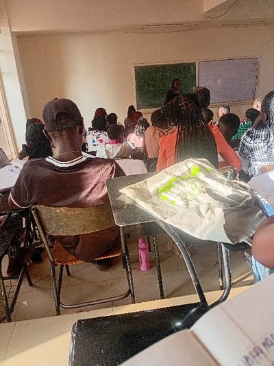 The image displays an indoor classroom in Karingani ward, Kenya, filled with approximately fifteen to twenty individuals, mostly students, attending an instructional session. In the foreground, facing away from the viewer, a male student with a dark baseball cap and a brown t-shirt is seated on a metal-framed chair with brown upholstery. To his immediate right, a female student with braided hair, wearing an orange top, sits at a desk that holds a light-colored item and several green writing implements. A pink water bottle is on the floor beneath a desk. Rows of students occupy similar metal-framed desks and chairs, facing a front wall where a female figure, presumed to be the lecturer, stands between a dark green chalkboard and a white board, both containing written information. Natural light enters the room from the left, illuminating parts of the scene. The walls are light-colored, and the floor is tiled.