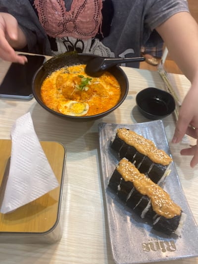 This image captures a close-up view of a table setting at what appears to be a restaurant in Watampone, Indonesia. Two individuals are present, with their hands visible, suggesting they are dining. In the foreground, on the right, a rectangular grey plate holds two servings of sushi rolls, each topped with a dollop of orange sauce and sprinkled with sesame seeds. To the left of the sushi, a white paper napkin is folded and placed on a wooden holder. In the background, a black bowl contains a serving of ramen or a similar noodle soup, garnished with chopped green onions and a slice of hard-boiled egg. A black spoon rests in the bowl. A small black dipping dish is positioned between the ramen and the sushi. A smartphone with a black screen is visible on the left side of the table, next to the ramen bowl. The table surface has a light-colored wood grain pattern. The overall lighting suggests an indoor setting, likely a casual dining establishment. The presence of chopsticks in a wrapper further indicates a meal is being served.