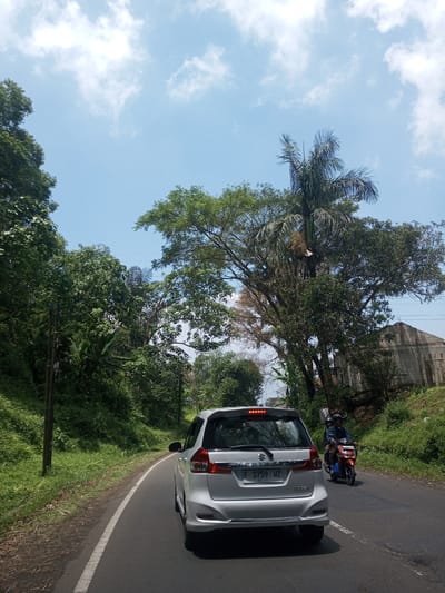 A silver Suzuki car is traveling on an asphalt road bordered by lush green vegetation and tall trees under a partially cloudy sky. A red and black motorcycle with two helmeted riders approaches from behind the car on the right side of the road. A partly-constructed concrete building is visible through the trees on the right. The car's license plate reads "F 1759 WZ". The specific location within Nawng Hkar, Myanmar cannot be determined from the given information.