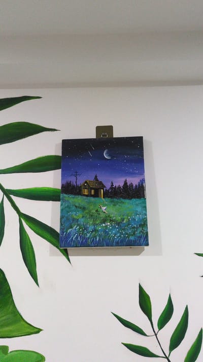 A square painting hangs on a white wall, held by a small, dark-colored clip. The painting depicts a landscape scene with a dark blue, star-filled night sky featuring a crescent moon and shooting stars. Below the sky, a line of dark green trees borders a field of green and blue grass with small white flowers. In the center of the painting, a house with lighted windows stands. A small white bird flies above the grass. The wall also has painted green leaves, resembling a plant. There is no available information about the image being located in the city of Phan Rang – Tháp Chàm, Vietnam.