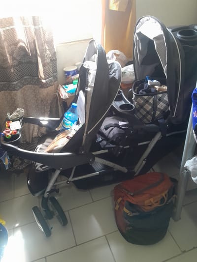 The image shows a twin stroller parked on a tiled floor inside a room. The stroller is laden with various items, including a bottle of water, food items, and a checkered storage basket filled with clothes. In the background, a window is partially covered by a patterned curtain, allowing sunlight to stream in. Beside the stroller, a backpack is placed on the floor. The scene appears to be in a residential setting, possibly a home, in Dam Camp, Nigeria. There are no people visible in the image. The overall impression is one of domestic clutter.