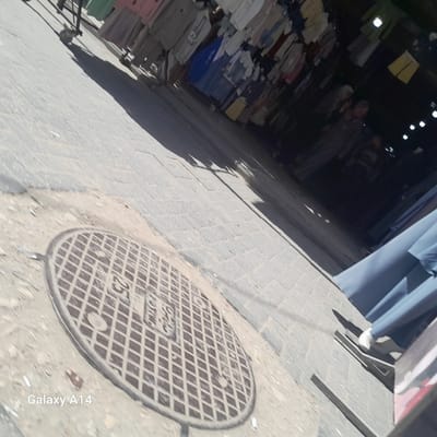 The image depicts a street scene in what appears to be the city of Nuseirat, Palestinian Territory, based on the context provided. The image has a strong diagonal perspective. A manhole cover is located in the lower left quadrant. The street is paved with light-colored bricks. Along the right side of the frame, there are pieces of clothing on a garment rack. In the background, more clothes appear to be displayed in a store. A person, who may be a shopkeeper, is standing in the background. In the far left corner, there is a glimpse of a garment rack with wheels.