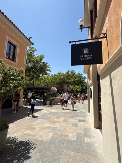 The image shows a street scene with several people walking. The architecture suggests a European setting, with buildings featuring stucco exteriors and tiled roofs. A sign with the text "LA ROCA VILLAGE" is prominently displayed, indicating the location. The day appears to be sunny, with clear blue skies and shadows cast by the buildings and trees, suggesting it is daytime, likely late morning or early afternoon. The people are dressed casually, and their expressions are not clearly discernible, but the overall atmosphere seems relaxed and leisurely, typical of a shopping district. One building has a decorative light fixture hanging from it. The ground is paved with large, irregular stone tiles, and there are trees and greenery interspersed throughout the scene, adding to the pleasant outdoor ambiance.